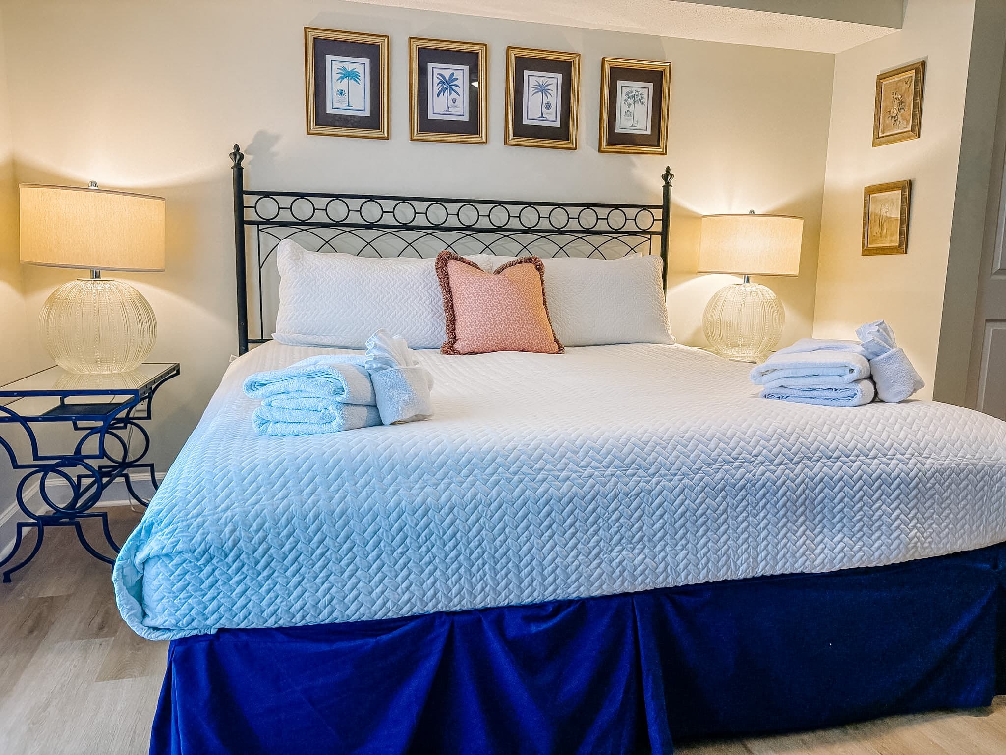 White quilted bed with blue skirt, pink pillow, and palm tree prints above the headboard.