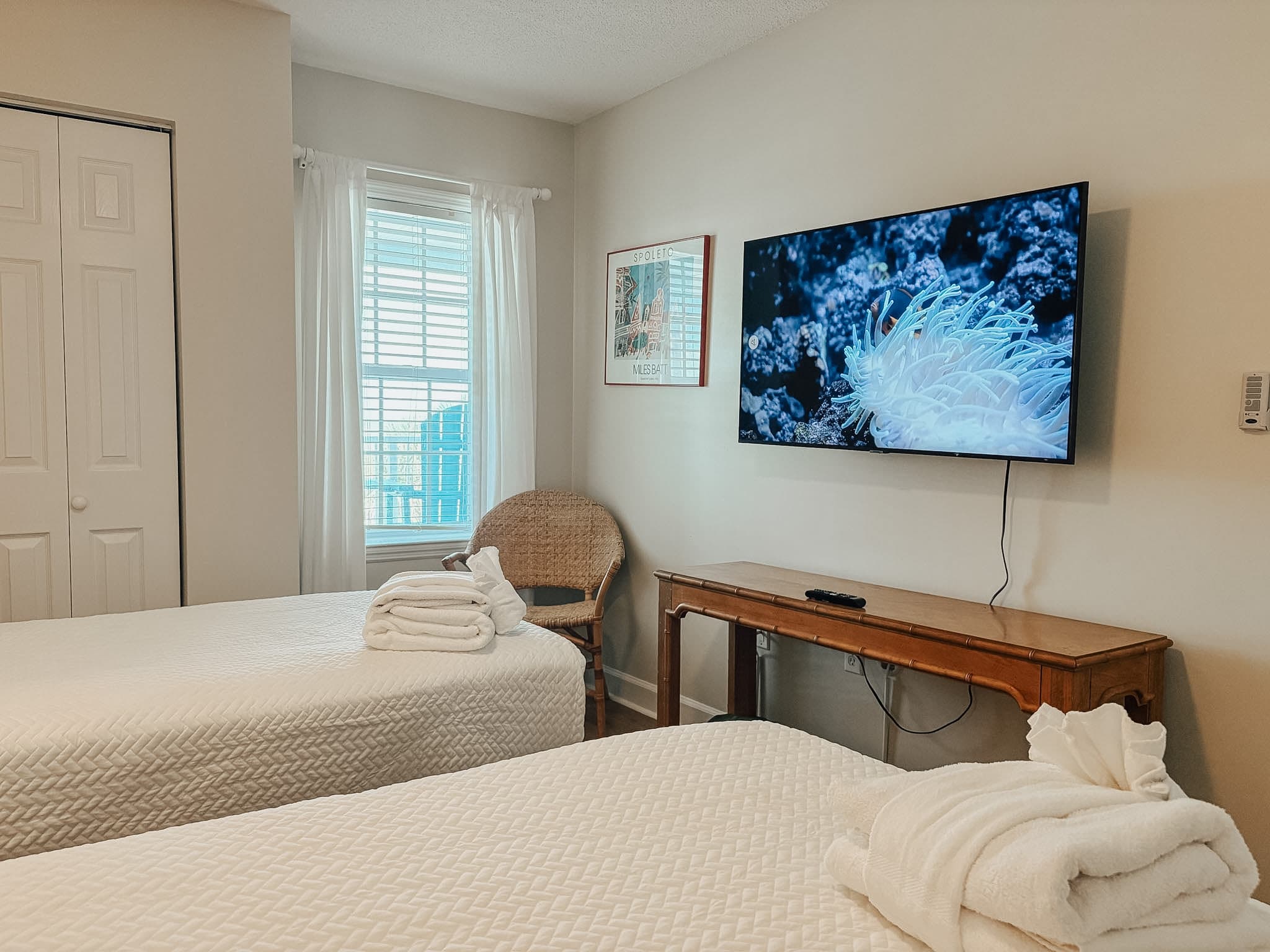 Bright bedroom featuring two twin beds, white towels, and a large wall-mounted television.