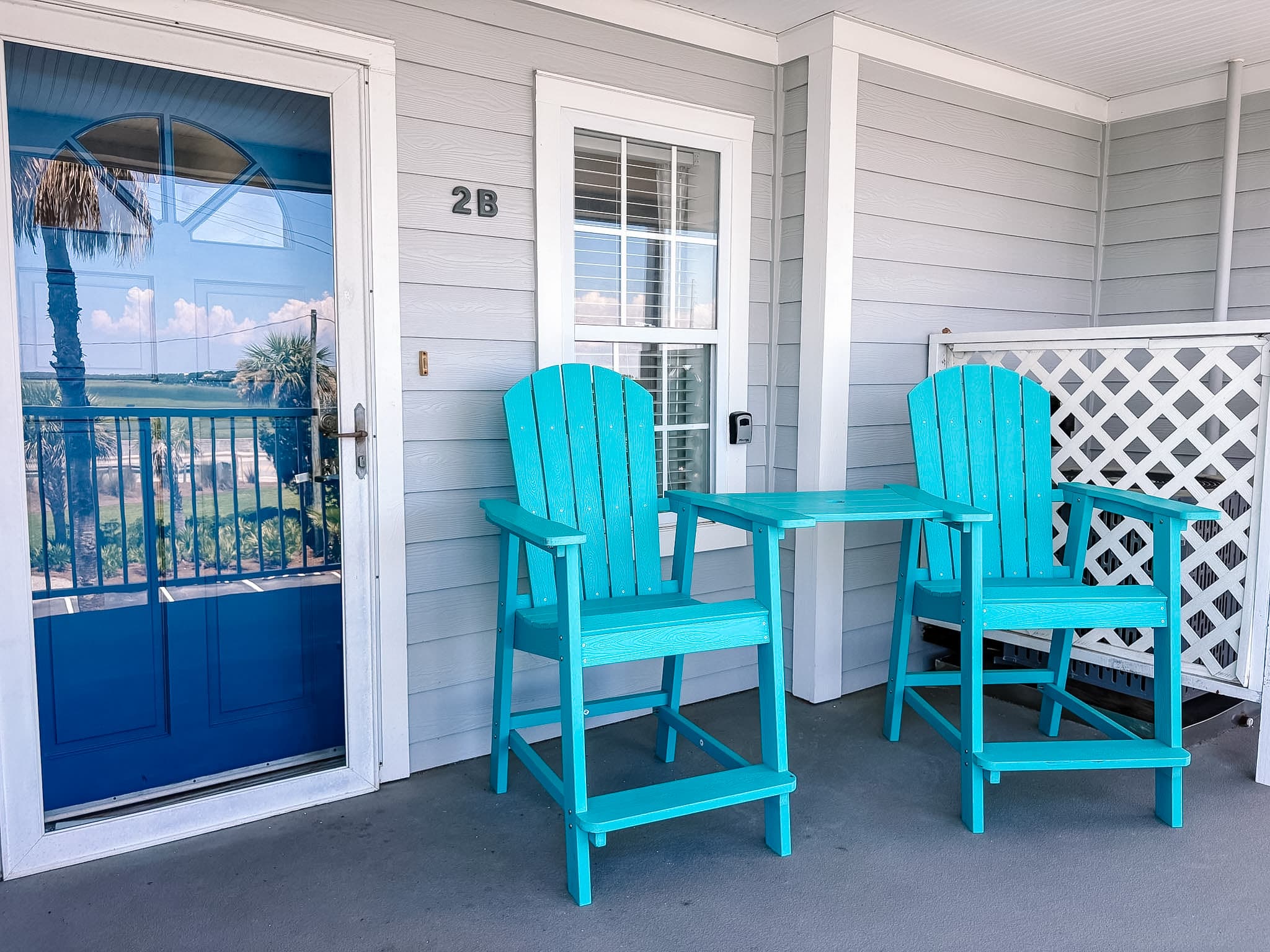 Bright turquoise Adirondack chairs and a small table on a porch beside a blue door.